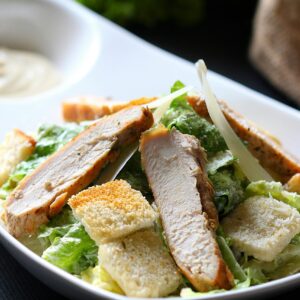 A close-up of a Caesar salad topped with grilled chicken slices, croutons, and dressing.