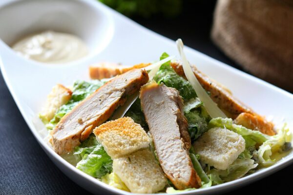 A close-up of a Caesar salad topped with grilled chicken slices, croutons, and dressing.